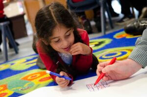 A young girl lying on her stomach, holding a pen and looking at a book.