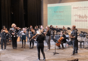 Pupils playing folk music with wood string instruments on stage, led by an instructor at the front.