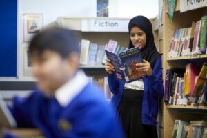 Pupils in the school library, a boy sits in the foreground, whilst a girl stands beside the bookshelf behind him, reading a book.