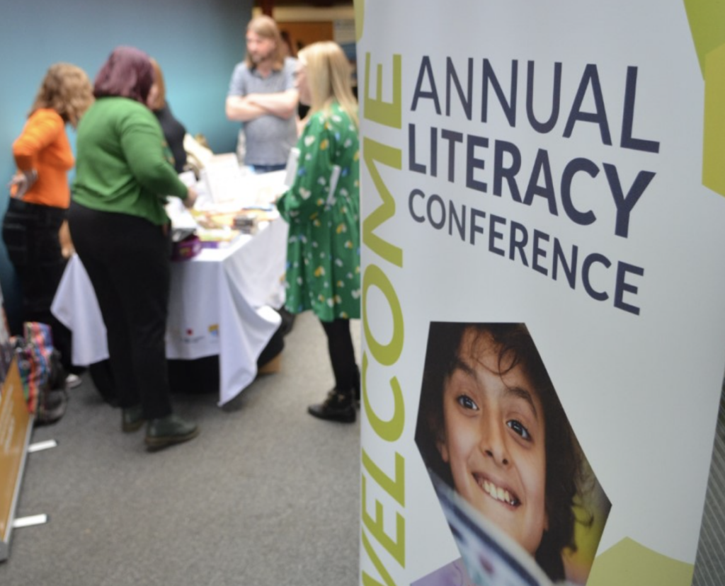 A welcome sign to the Literacy Conference, with delegates talking to each other in the background.