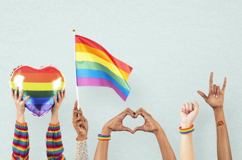 Hands raised in the air, holding a rainbow heart, a rainbow flag, and wearing rainbow wristbands.