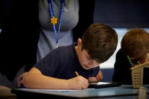 A boy writing at his desk with a teacher nearby.