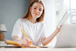 A woman sat at a desk, holding a book in one hand and writing with the other.