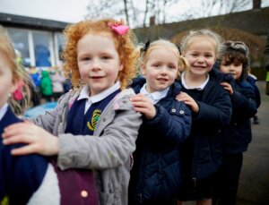 A group of young children in the playground, playing follow the leader with their hands on each others shoulders.