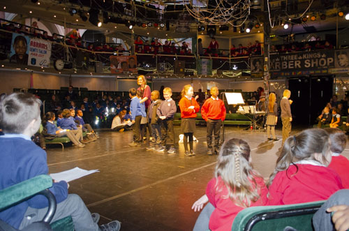 Children stand under the spotlights on stage, whilst others sit in the audience. 