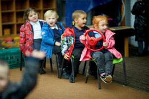 Four young children in the early years, sitting on chairs behind each other to create a bus, whilst the child at the front holds a steering wheel.